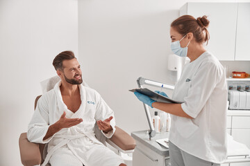 Obraz premium Cosmetologist in mask and gloves enterviews bearded man before procedures in beauty salon