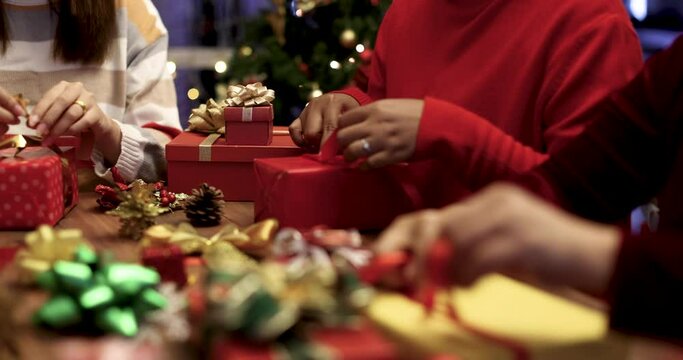 Hands of three women in red winter dress holding and using ribbon to tie bow for a wrapped gift box for Christmas present with Xmas tree in front and colorful lights bokeh in blur background.