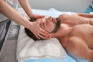 Fototapeta na wymiar Massage therapist works with head of male lient lying on couch in contemporary clinic