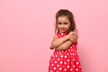 Beautiful preschool 4 years kid girl shyly hugging herself, cute smiling posing on camera against pink wall background with copy space. Confident portrait of shyly gorgeous baby girl. Adorable child