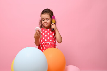Obraz na płótnie Canvas Isolated portrait on pink background of a gorgeous baby girl in stylish pink dress, standing behind multicolored balloons, combing her curly hair , looking at her reflection in small cosmetic mirror.