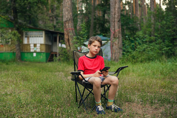 boy sitting in a folding chair and holding a smartphone against the background of summer nature