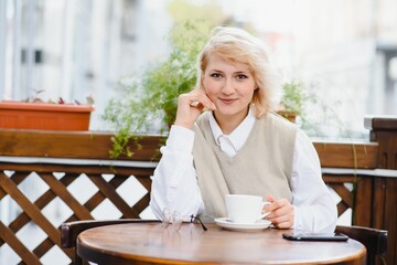 young stylish beautiful woman sitting in city cafe in street, spring fashion trend style, drinking latte, pink purse, exited, emotional, happy, smiling