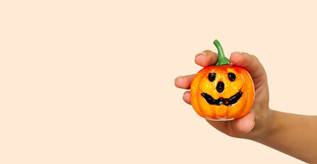 A young girl's hand holds a small orange pumpkin for Halloween against the background of the color trend #FAEBD7 Champagne 2021. A smiling Jack lantern is a symbol of the Halloween holiday. Horror