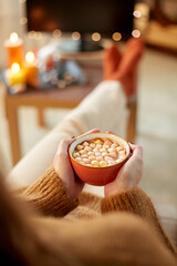 halloween, holidays and leisure concept - close up of young woman watching tv and drinking hot chocolate with marshmallow with her feet on table at cozy home