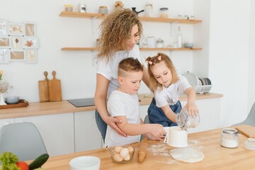 Happy young mother with cute little preschooler kids have fun making dough baking pie or pastry in modern kitchen together, overjoyed mother teach small children doing bakery cooking at home