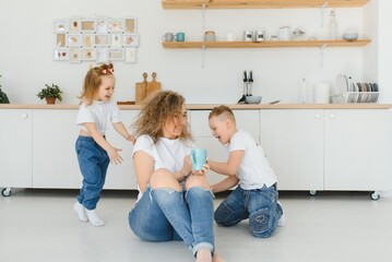 Happy mother hugging daughter and son sitting on wooden floor in modern kitchen at home. Mom hugging children. Happy family relationship concept