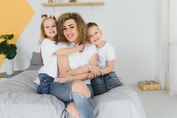 Obraz premium Portrait of smiling young mommy embracing kids little son and daughter. Happy family. Mother with children sitting on bed and loving the good times together. Closeup