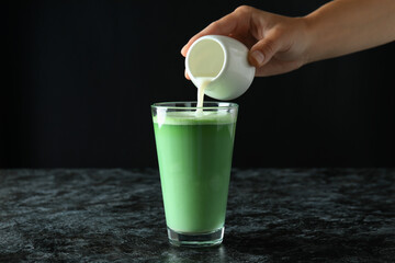 Female hand pouring milk into a glass of matcha latte against black background