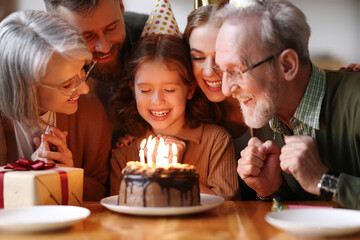 Cute excited little child making wish, blowing candles on cake while celebrating Birthday with family