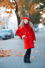Stylish happy kid girl 5-6 year old with blonde curly hair wear red color jacket and beret hat walk in city street. Autumn season. Childhood. Looking at camera.
