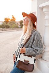 Stylish blonde young adult girl 22-24 year old wear felt hat, denim pants and knit woolen cardigan...