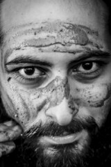 Tight black and white portrait of a bearded middle eastern man putting mud on his face