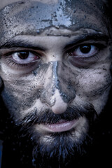 Tight portrait of a bearded middle eastern man with the face covered by mud and an intense gaze	