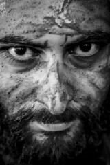 Tight black and white portrait of a bearded middle eastern man with the face covered by mud and an intense gaze