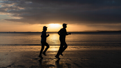 Silhouette of two people running on sand beach at sunrise