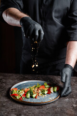 The hand of a professional chef with a black glove sprinkling spices on a prepared salad. Background for a restaurant or for a menu with space for your text