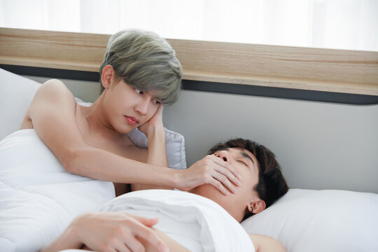 Lgbt gay couple having problems in bed. Young man with sullen face covering his ears and using hand close his boyfriend's mouth who snoring loudly disturbing his sleep