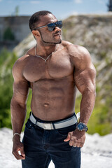 Strong stylish man wearing glasses posing on clear sandy beach. Shirtless bodybuilder showing his...