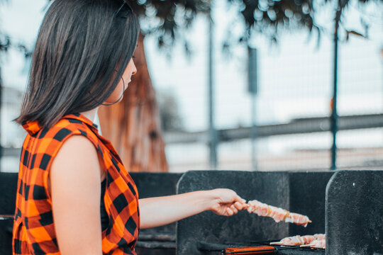 Chica joven de camisa de cuadros y morena cocinando varias comidas en una barbacoa junto a los arboles