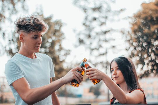 Chica y chico joven brindando con cervezas en el parque al aire libre junto a los arboles