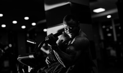 Fototapeta na wymiar Male afroamerican athlete is doing biceps dumbbells curls while sitting in gym on sport chair. Black and white photo