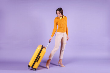 Tourist woman wearing mustard sweater, beige pants and yellow earphones is carrying yellow suitcase...