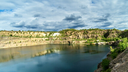 Fototapeta na wymiar Panoramic view of an old flooded granite quarry against cloudy sky in early summer. Landscape with rock stones, green trees and clean pond