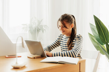 A dark-haired schoolgirl sits at a desk doing her homework using her tablet. Modern technologies, online education, training