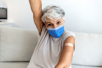 Mature woman against white background after receiving COVID-19 vaccination, wearing protective face mask. Old Caucasian lady holding up shirt sleeve to show the sticking plaster after a flu jab in arm - obrazy, fototapety, plakaty