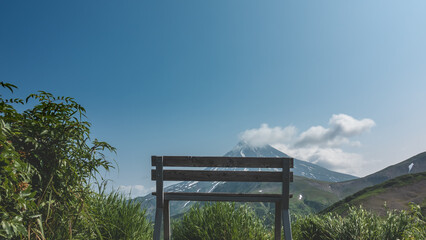 There is an unpainted wooden bench among the green grass. Ahead, against the blue sky, a conical volcano. There is snow on the slopes. Clouds over the top. Kamchatka. Stratovolcano of Vilyuchinsky