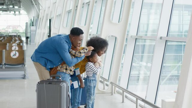 Multiracial father with suitcases on wheels and little kids standing near the big glass window in the airport and looking outside. Traveling concept