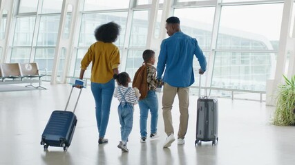 Back view on the multiracial family - mother, father, daughter and son - walking through the airport hall and carrying their suitcases on the wheels before the departure to the vacation - Powered by Adobe