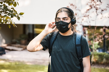 Young teenager high school boy with headphones and backpack wearing medical mask and listens to music at the school garden.