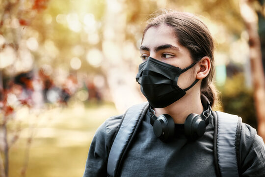 Concerned young teenager high school boy with headphones and backpack wearing medical mask at the school garden. - Powered by Adobe