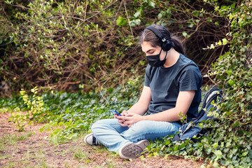 Teenage high school boy with medical mask sitting in cross-legged position at the school garden and texting with his smartphone