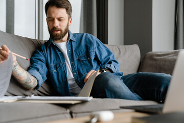 Fototapeta premium Bearded european man working with papers and laptop while sitting on sofa