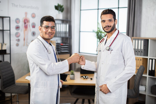 Experienced two male Indian Asian doctors surgeons in medical coats making fists bump, standing in modern hospital office, preparing for operation. Team of two doctors bumping fists