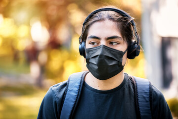 Concerned teenager high school boy with headphones and backpack wearing medical mask at the school garden.