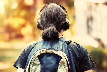 Facing back teenager high school boy with headphones and backpack walks towards the school building.