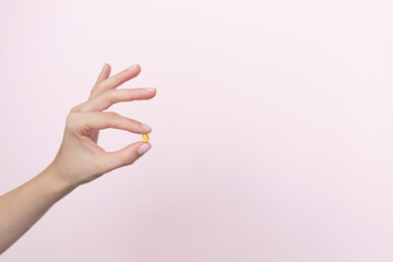 Woman hand holding yellow pill capsule on a pink background, medication concept with copy space