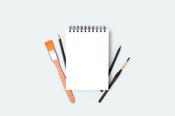 Empty notepad mockup and tools for painting on a blue pastel background. Notebook, paintbrush and pencil with copyspace. Artistic workspace.
