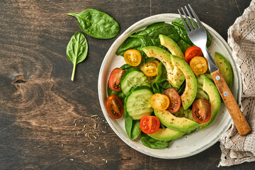Avocado, red, yellow, black cherry tomato, spinach and cucumber fresh salad with spices pepper and olive oil in grey bowl old wooden table background. Healthy food concept. Top view.