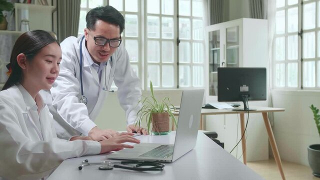 Hospital Medical Doctor'S Office: Two Asian Professional Physicians Talking, Consulting And Working On Laptop Computer. Specialists Discuss Patient Online Treatment, Writing Digital Medicine Prescript