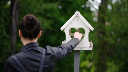 feeds birds in a feeder in an spring park. girl puts food. girl out of focus, rear view. taking care of birds, natural green blurred background. wooden birdhouse. white feeder in the garden. close-up