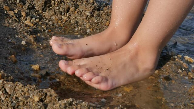 the bare feet of a child stomp in puddles of water on the sea sand with pebbles. Slow motion, close-up.