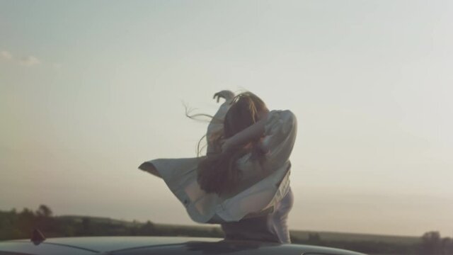 Young beautiful girl rides in the car with raised hands up, a woman in the sunroof of the car. Enjoying life at sunset in a car hatch. Hair in the wind in slow motion. Road trip adventure, wanderlust