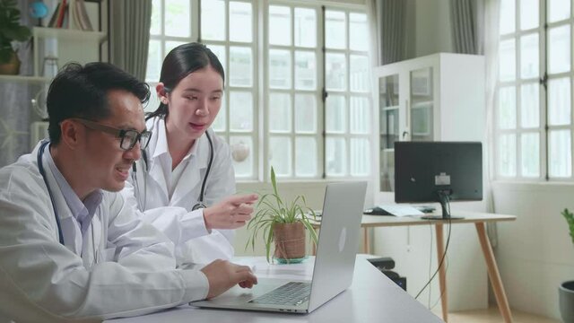 Hospital Medical Doctor'S Office: Two Asian Professional Physicians Talking, Consulting And Working On Laptop Computer. Specialists Discuss Patient Online Treatment, Writing Digital Medicine Prescript