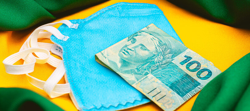  Real - BRL. A pandemia do novo coronavírus e economia brasileira. Uma cédula de 100 reais sobre uma máscara n95 de cor azul. Na composição da foto uma representação da bandeira do Brasil.