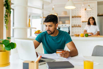 A young African American man sits at the dining table and shopping online a git for his wife. A man is holding a credit card and searching perfect gift while the woman is preparing dinner. - 451601174